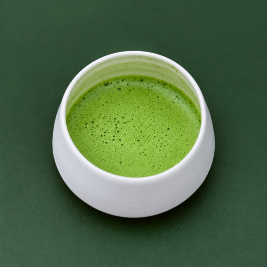 Taça branca com matcha cerimonial verde batido e espuma fina, sobre fundo verde.